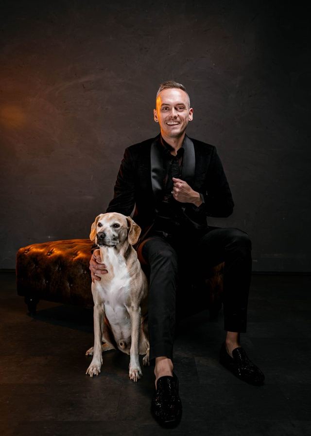 Tyler smiling in a black velvet jacket, seated beside Banksy, his tan mixed-breed dog, in a studio