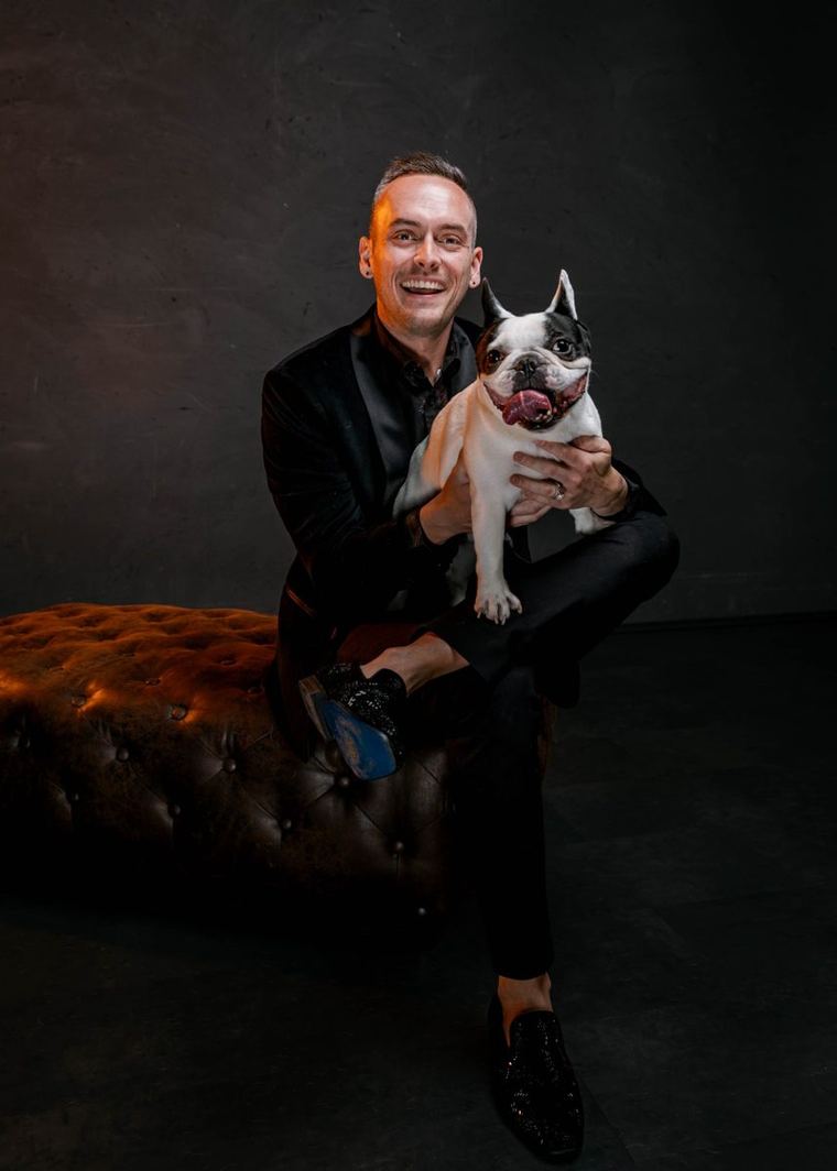Tyler laughing in a studio, sitting on a leather ottoman with Leo, a black and white French Bulldog, in his arms